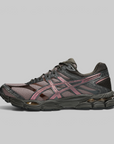 A single ASICS running shoe in a dark charcoal gray and black mesh with metallic rose gold overlays, shown in a side profile on a solid gray background.