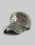 A baseball cap in a green and brown camouflage pattern, featuring a white embroidered 'LA' logo on the front and a small white '47' brand logo on the side, set against a solid gray background.
