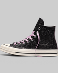 A high-top sneaker in black embossed leather with light pink laces, a cream rubber sole, and a small silver logo on the side, shown against a solid gray background.
