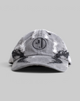 A gray washed baseball cap with distressed details, black embroidered leaf patterns on the sides, and a circular black monogram logo on the front, set against a solid gray background.