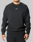A man wearing a black Nike crewneck sweatshirt with a white swoosh logo on the chest and 'Mood' text on the sleeve, paired with black pants, set against a solid gray background.