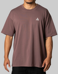 A man wearing a muted mauve-brown short-sleeve t-shirt with a small white geometric triangle logo on the left chest, paired with black pants, against a solid gray background.