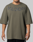 A man wearing an oversized olive green t-shirt with a dark gray mirrored 'NOCTA' logo printed across the chest, shown against a solid gray background.