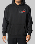 A man wearing a black pullover hoodie with a small chest graphic showing a red heart and a gray-and-white skeletal hand, photographed against a plain light gray studio background.