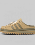 A low-top slip-on sneaker in beige suede with olive green stripes and laces, featuring a chunky gum sole with a zigzag tread, shown against a solid gray background.