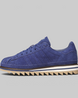A single blue suede sneaker with a chunky, jagged gum sole, photographed from the side against a solid light gray background.