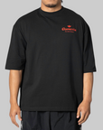 A man wearing an oversized black t-shirt with a red 'Chavarria' logo printed on the left chest, photographed against a solid gray background.
