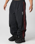 Black track pants with red side stripes and a small white logo, shown on a model from the waist down against a light gray studio background.