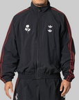 A man wearing a black zip-up track jacket with red stripes down the sleeves and white embroidered chest graphics, paired with matching black track pants, against a solid gray background.