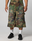 A person wearing knee-length camouflage cargo shorts with large front flap pockets, shown from the waist down against a light gray studio background. The shorts feature a woodland camo pattern in green, brown, black, and tan, with a small embroidered chest logo.