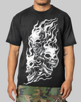 A man wearing a black short-sleeve t-shirt with a large white graphic of three skulls surrounded by flames, paired with camouflage pants, against a solid gray background.