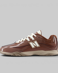 A New Balance low-top sneaker in brown patent leather and mesh, featuring white laces and a large white 'N' logo with a gold outline on the side, set against a solid gray background.