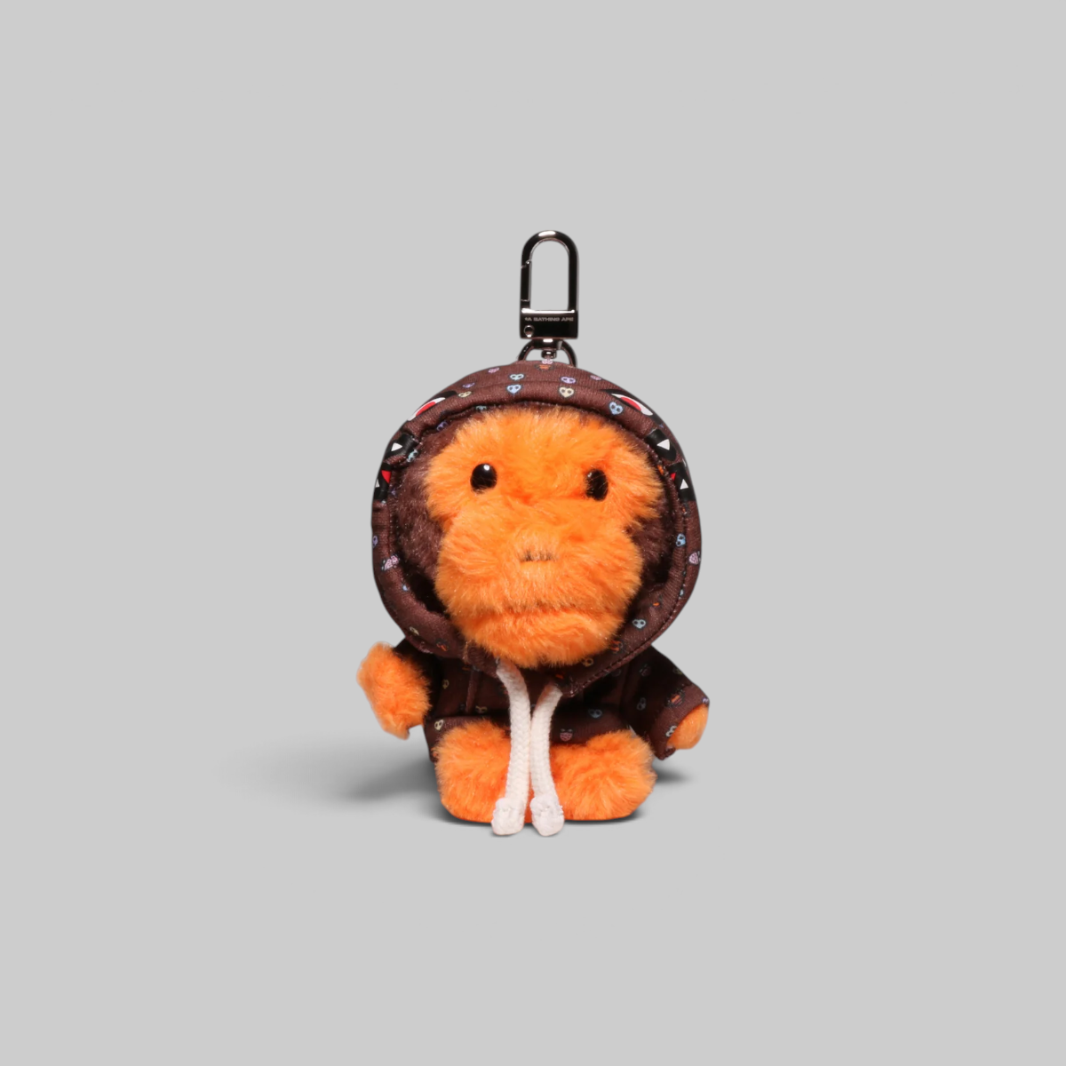 A plush keychain or bag charm shaped like a small orange creature wearing a brown hooded outfit with small colorful dot details, shown on a plain light gray background.