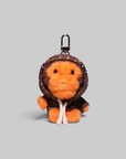 A plush keychain or bag charm shaped like a small orange creature wearing a brown hooded outfit with small colorful dot details, shown on a plain light gray background.