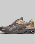 An ASICS sneaker in dark brown and tan, featuring the brand's signature side stripe design and a dark gray cushioned sole, shown against a solid gray background.