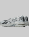 A pair of metallic silver athletic sneakers with mesh panels and reflective synthetic overlays, shown from a side profile view against a solid gray background.
