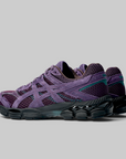 A pair of ASICS running shoes in purple and black, featuring the brand's signature side stripe design, shown against a neutral gray background.