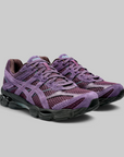 A pair of Asics running shoes in a purple and burgundy mesh and suede upper, with black and gray cushioned soles, photographed on a solid gray background.
