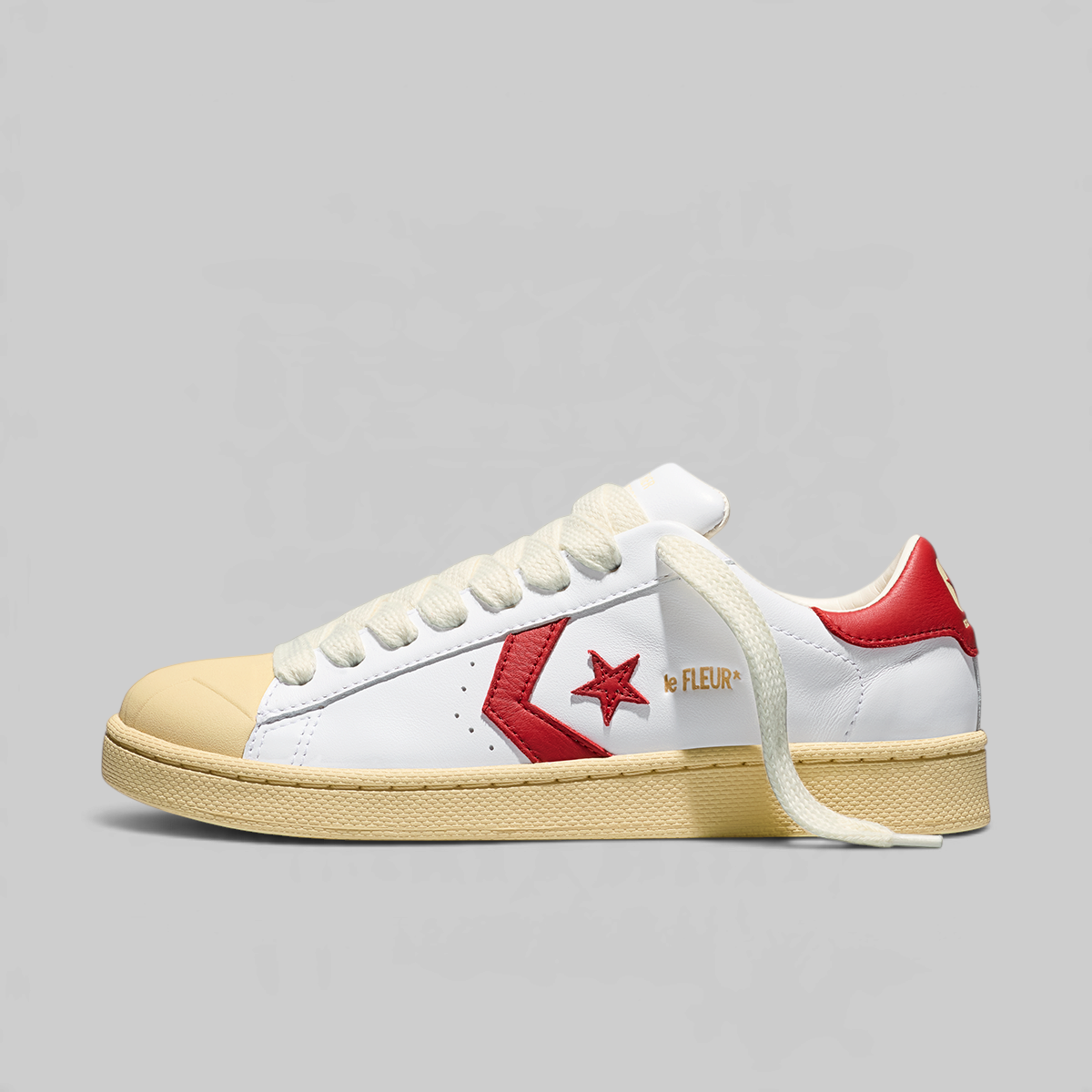 A Converse sneaker in white leather with a red star logo, red heel tab, gold 'CONVERSE' text on the side, a beige rubber toe cap and midsole, and white laces, photographed on a light gray background.