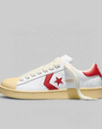 A Converse sneaker in white leather with a red star logo, red heel tab, gold 'CONVERSE' text on the side, a beige rubber toe cap and midsole, and white laces, photographed on a light gray background.