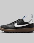 A Nike low-top sneaker in black textured leather with a white Swoosh, a thick brown gum sole, and blue pull tabs, shown in a side profile on a light gray background.