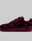 A single low-top sneaker in a deep burgundy faux fur material, shown from a side profile against a solid light gray background.