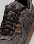 Close-up of a Nike sneaker in dark gray and brown suede with black laces and a translucent gum sole, showing the side swoosh and Nike branding.