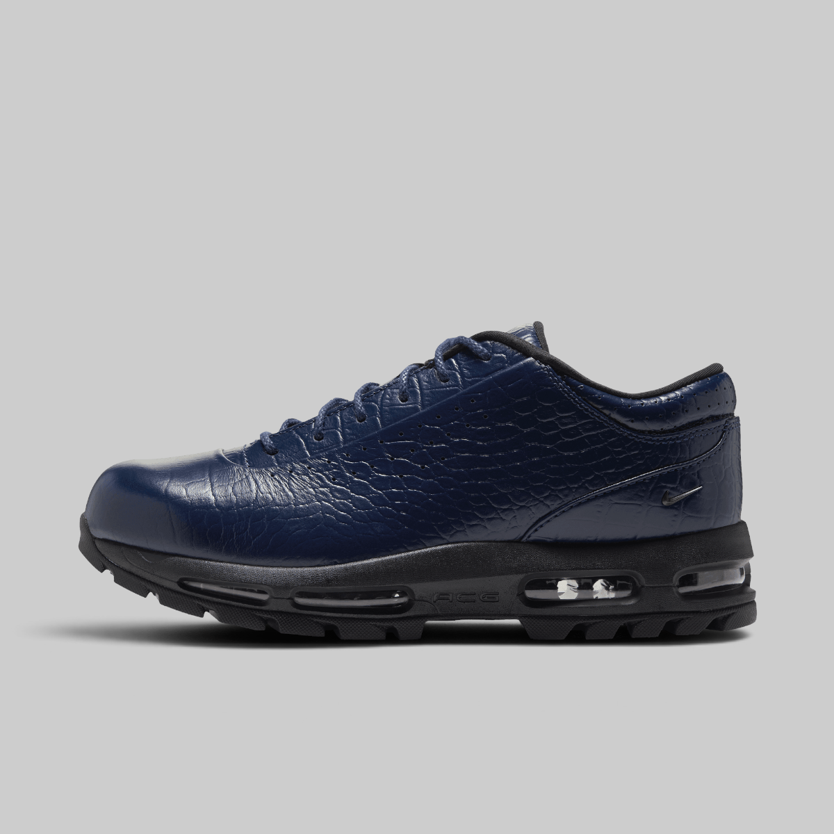 A navy blue Nike sneaker with a crocodile-embossed leather upper, black laces, and a black sole with visible air cushioning units, shown against a solid gray background.