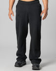 A person wearing black sweatpants and black sneakers, shown from the waist down against a light gray studio background.