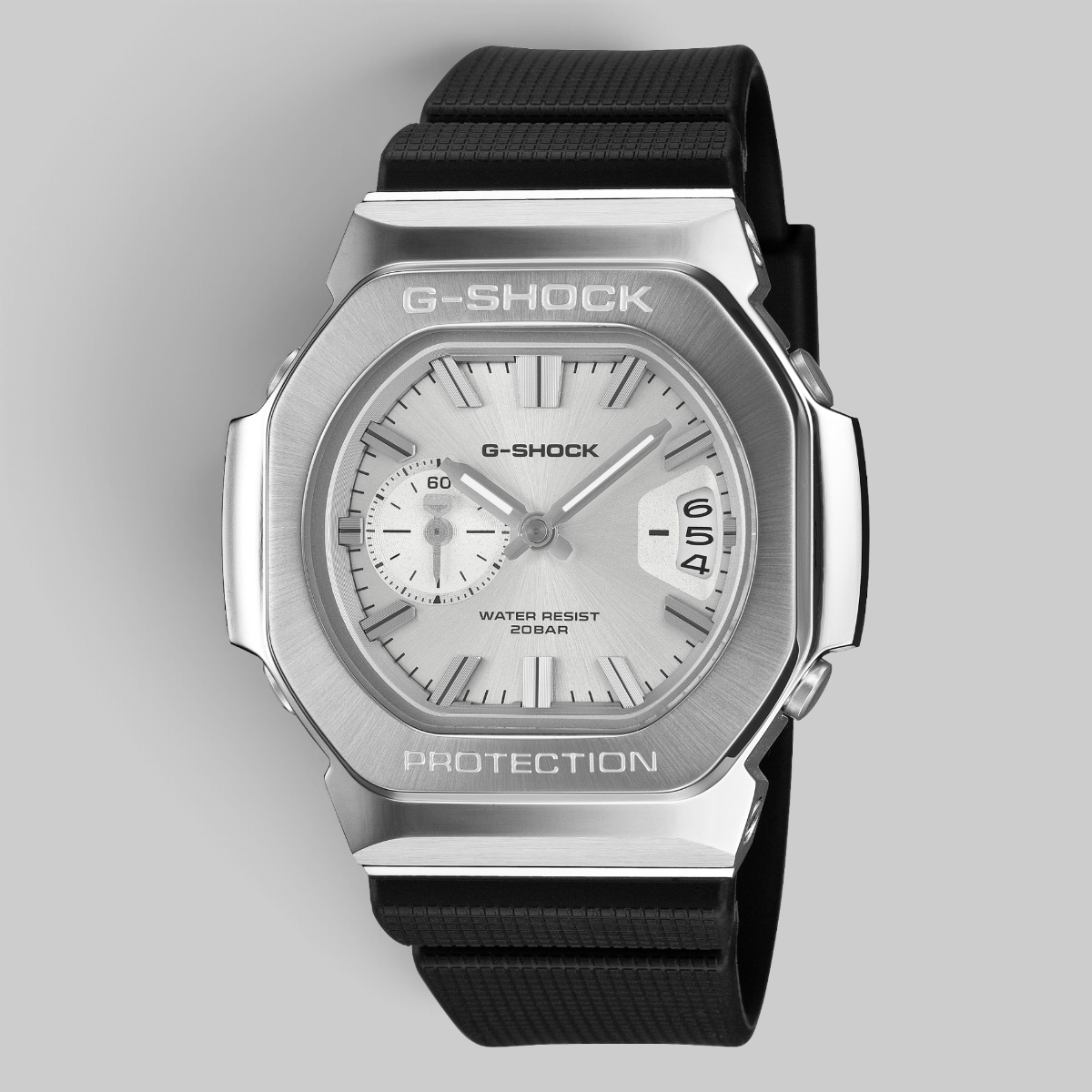 A G-Shock watch with a brushed silver-tone metal case, white dial, and black textured strap, shown on a neutral gray background.