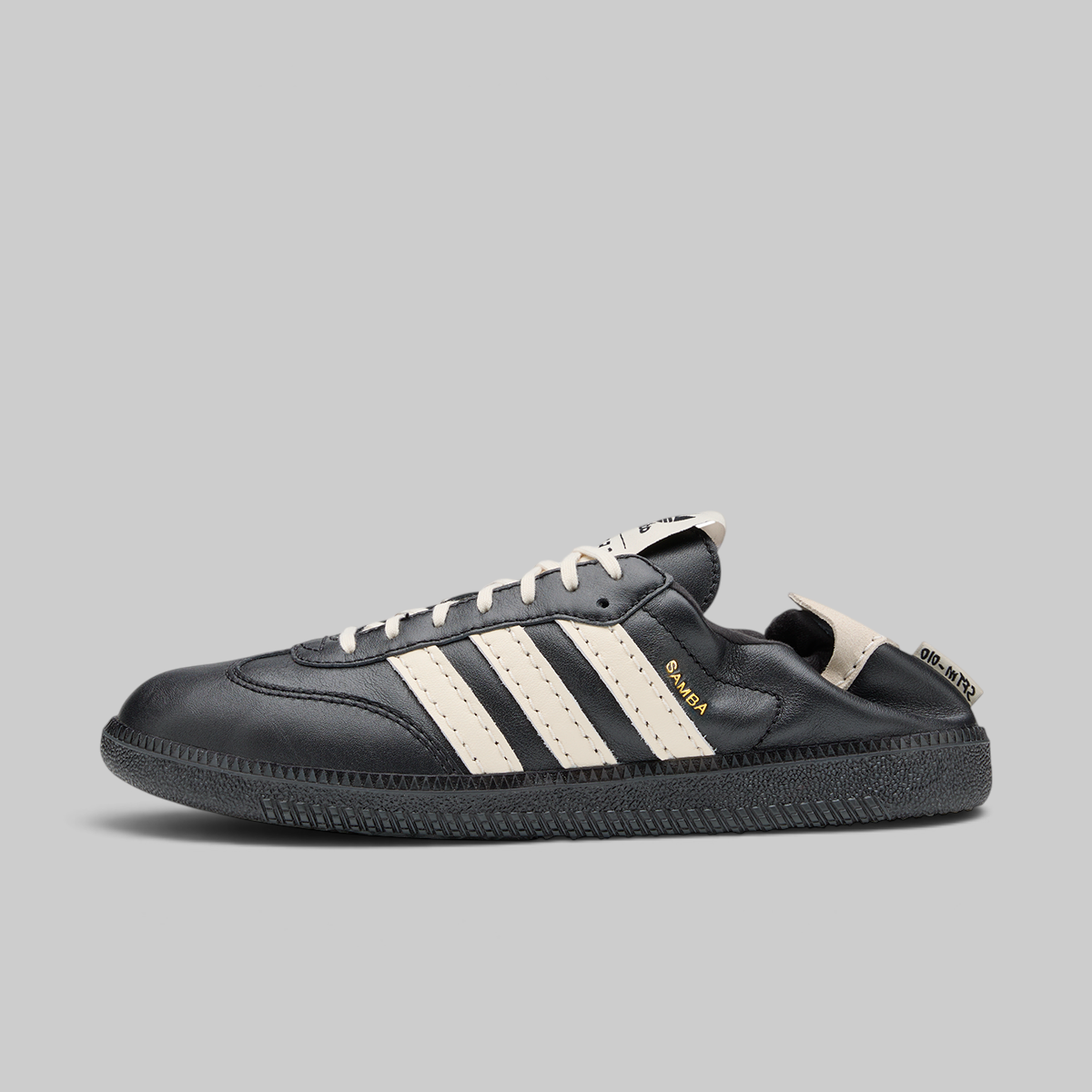 A black leather Adidas Samba sneaker with off-white three stripes, off-white laces, a black sole, and a small gold 'SAMBA' text on the side, shown against a solid gray background.