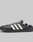 A black leather Adidas Samba sneaker with off-white three stripes, off-white laces, a black sole, and a small gold 'SAMBA' text on the side, shown against a solid gray background.