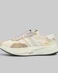 A low-top sneaker in cream and beige suede and mesh, featuring three white stripes on the side, a textured beige heel counter, a white rubber heel tab, and a chunky off-white platform sole with a black outsole.
