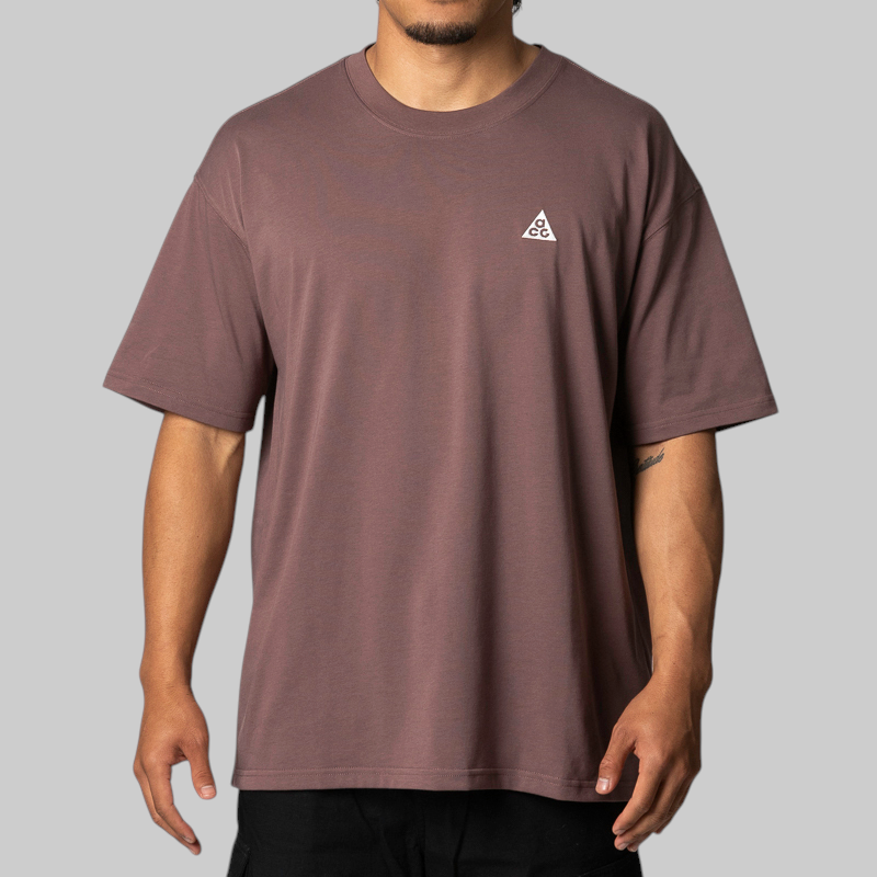 A man wearing a muted mauve-brown short-sleeve t-shirt with a small white geometric triangle logo on the left chest, paired with black pants, against a solid gray background.