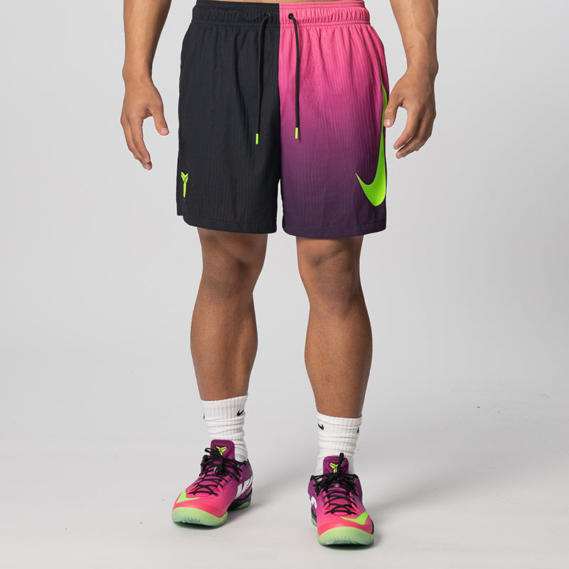 A model from the waist down wearing Nike athletic shorts with a split black and pink-to-purple design, neon green swoosh and drawstring tips, white socks, and matching pink and green sneakers against a light gray studio background.