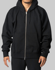 A man wearing a black Nike zip-up hoodie with a silver zipper, black drawstrings, and a small black swoosh logo on the left chest, set against a solid gray background.