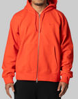 A man wearing a bright orange Nike full-zip hoodie with a silver zipper, black pants, and a beard, photographed against a solid gray background.