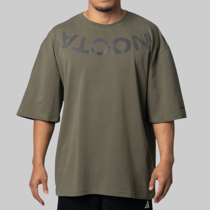 A man wearing an oversized olive green t-shirt with a dark gray mirrored 'NOCTA' logo printed across the chest, shown against a solid gray background.