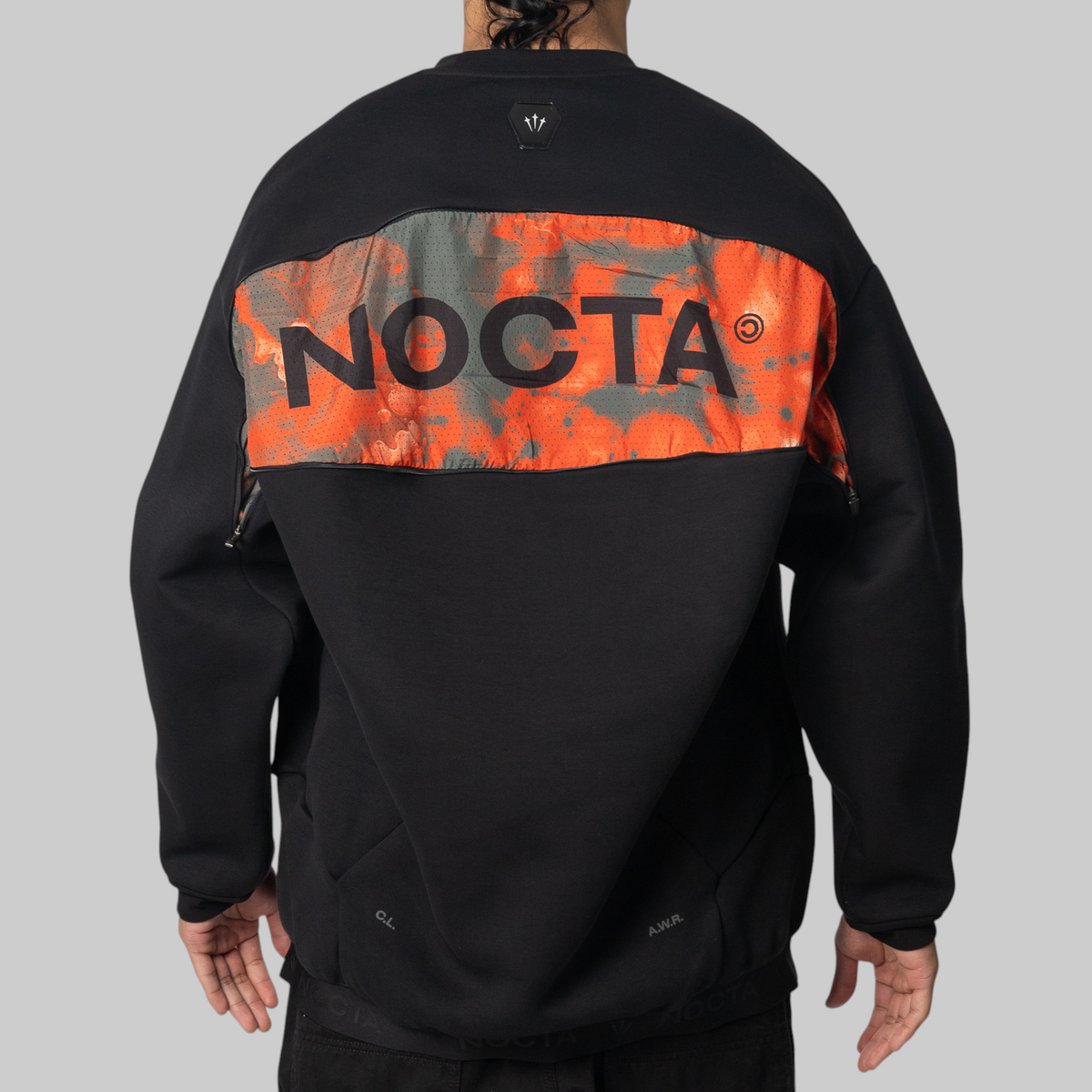 A person shown from the back wearing a black pullover sweatshirt with a wide orange and gray camouflage panel across the upper back featuring the word NOCTA in large black letters.