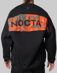 A person shown from the back wearing a black pullover sweatshirt with a wide orange and gray camouflage panel across the upper back featuring the word NOCTA in large black letters.
