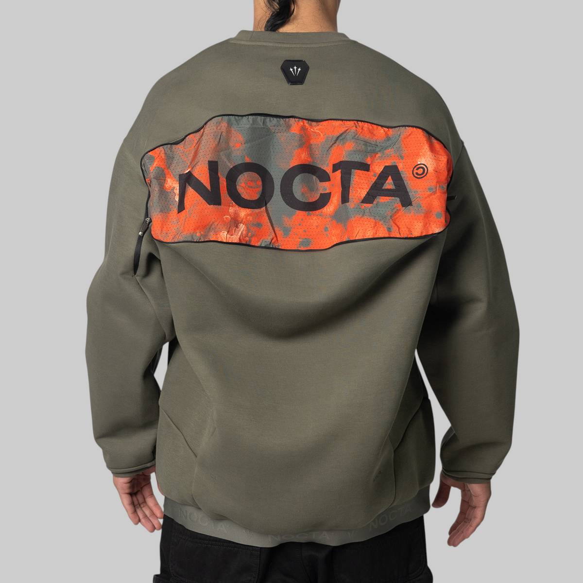 Back view of a person wearing an olive green pullover sweatshirt with a large orange and gray graphic panel across the upper back featuring the word NOCTA in black.