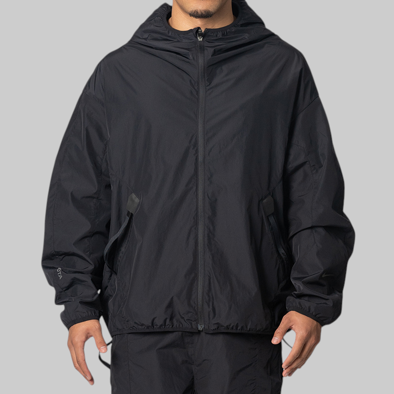 A man wearing a black hooded zip-up jacket and matching black pants, shown from the chin down against a plain light gray studio background.
