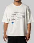 A man wearing an oversized off-white graphic t-shirt with black handwritten-style text and a small blue graphic on the front, paired with black pants, against a solid gray background.