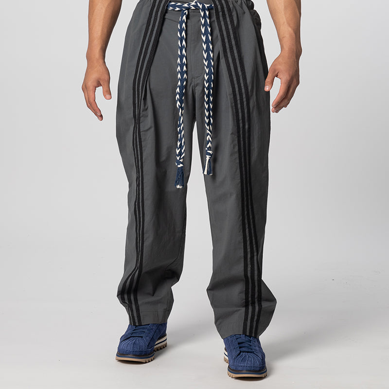 A person wearing dark gray baggy pants with black side stripes, a blue and white braided drawstring, and blue sneakers, shown from the waist down on a light gray studio background.