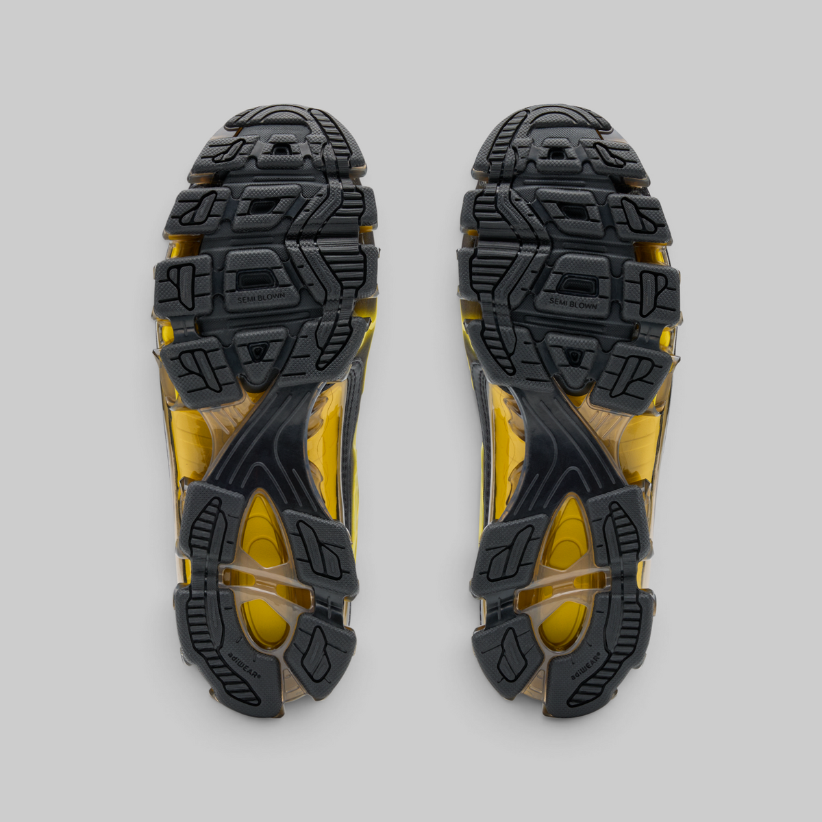Top-down view of a pair of athletic shoes showing their black and yellow textured outsoles against a solid gray background.
