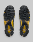 Top-down view of a pair of athletic shoes showing their black and yellow textured outsoles against a solid gray background.