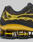 Close-up of a black and yellow athletic sneaker showing the side panel with a yellow lightning-bolt graphic, the translucent yellow midsole, and the black and yellow Shox sole.