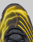 Close-up of a black athletic shoe with bright yellow lightning-bolt style graphics on the upper, shown against a light gray background.