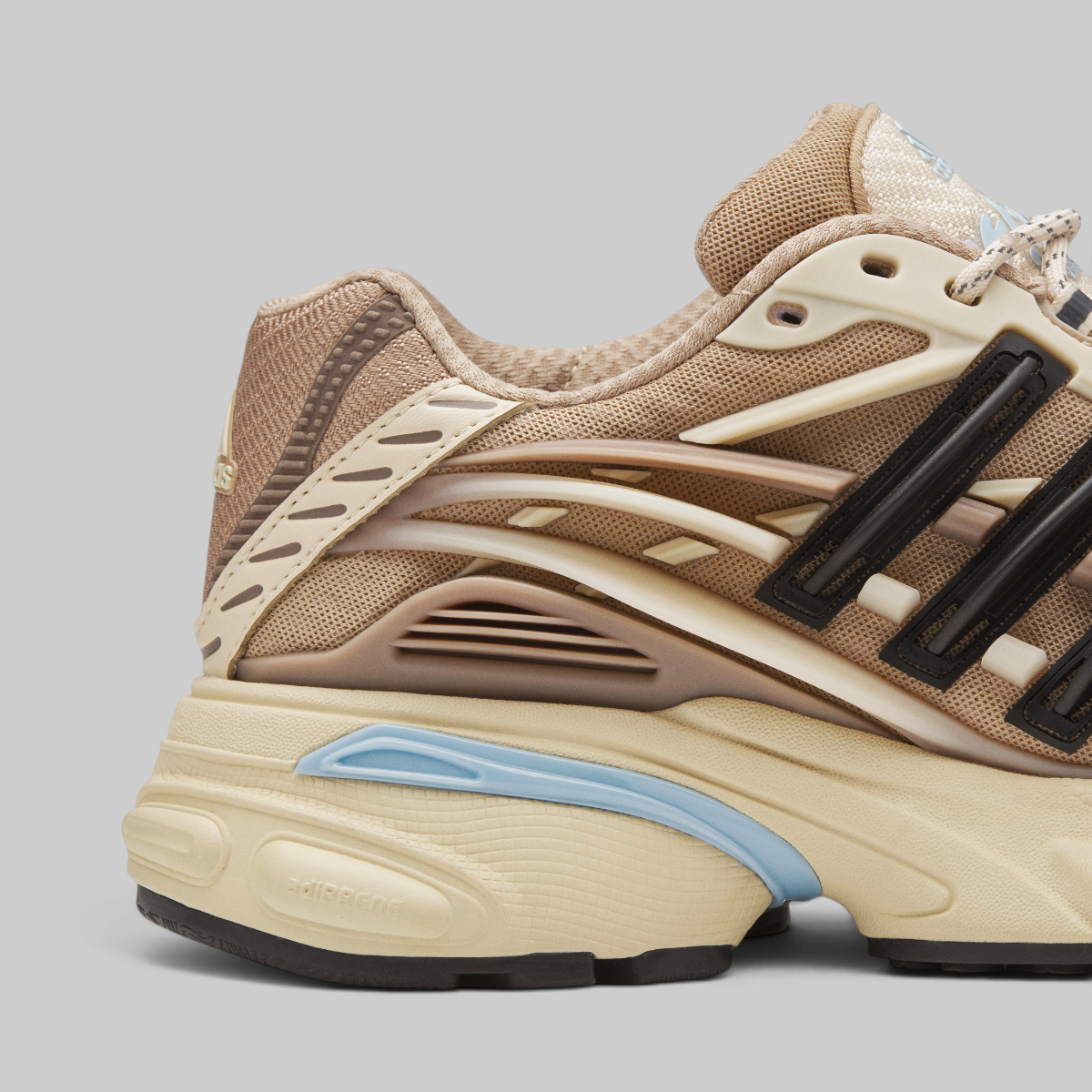 A close-up side profile of Pharrell Williams Adistar Jellyfish - "Crystal Sand" a chunky sneaker in beige and cream with black side stripes and a light blue accent on the midsole, set against a solid gray background.