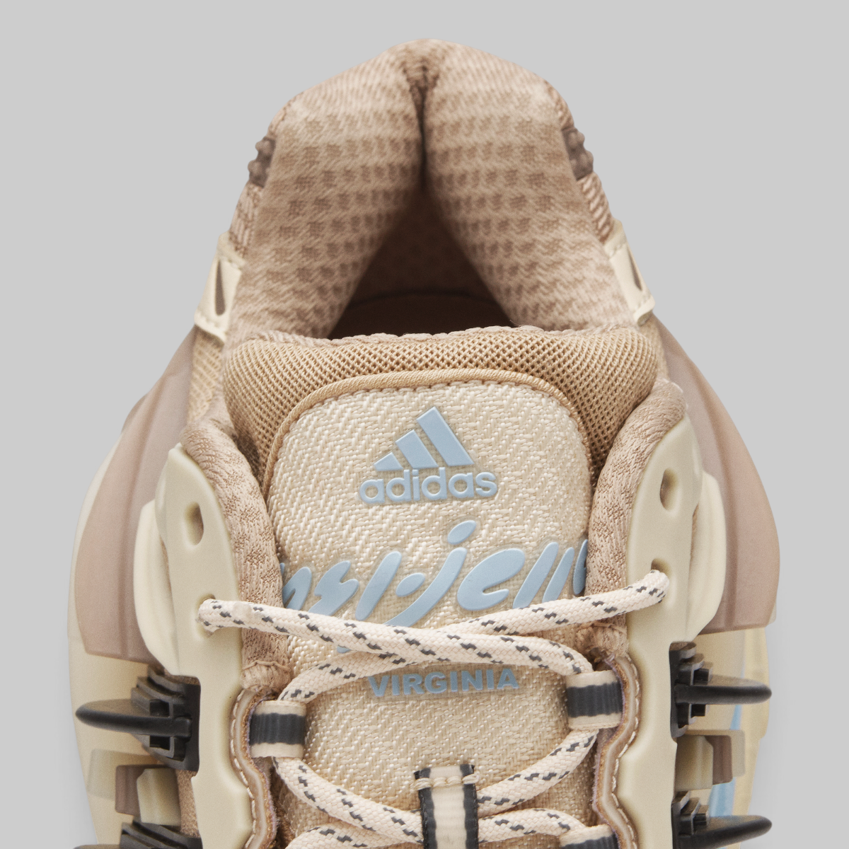 Close-up of the tongue and upper area of an Adidas Pharrell Williams Adistar Jellyfish - "Crystal Sand" sneaker in beige and cream tones, with light blue adidas branding and 'VIRGINIA' text on the lace guard, set against a solid gray background.